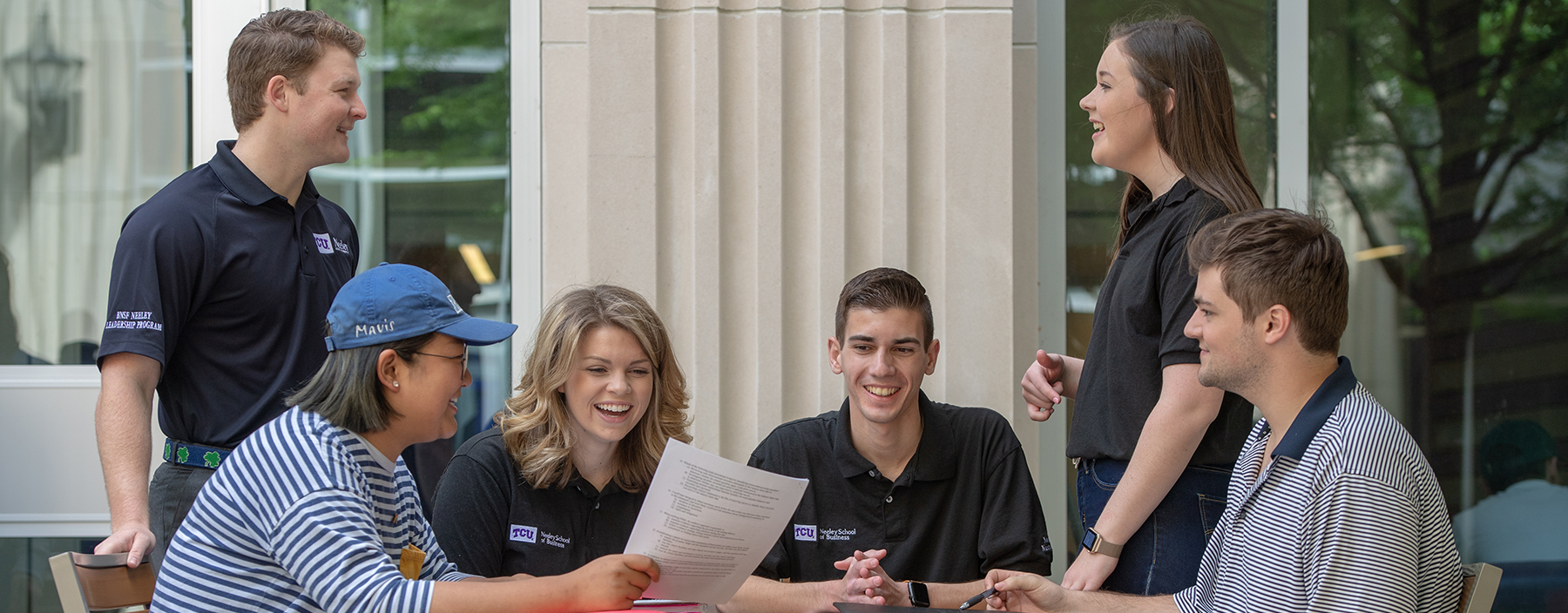 Section Image: Students gathered around a table outside, smiling and looking at papers. 