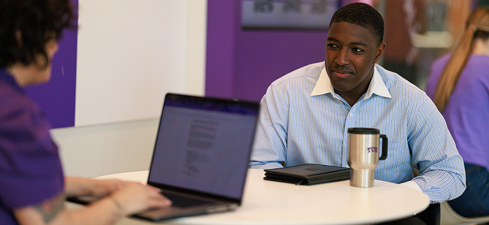 Man at a table looking at a man with an open laptop