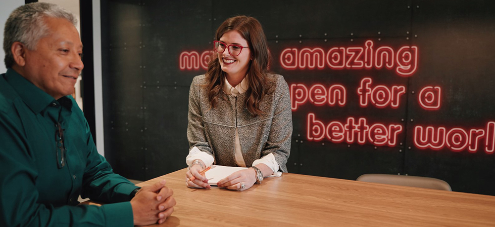 Section Image: Man and woman sitting at table with the words "make amazing happen for a better world" on the wall behind them. 