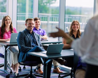 Smiling students with laptops listen as an instructor gestures during a class in a bright classroom with floor-to-ceiling windows