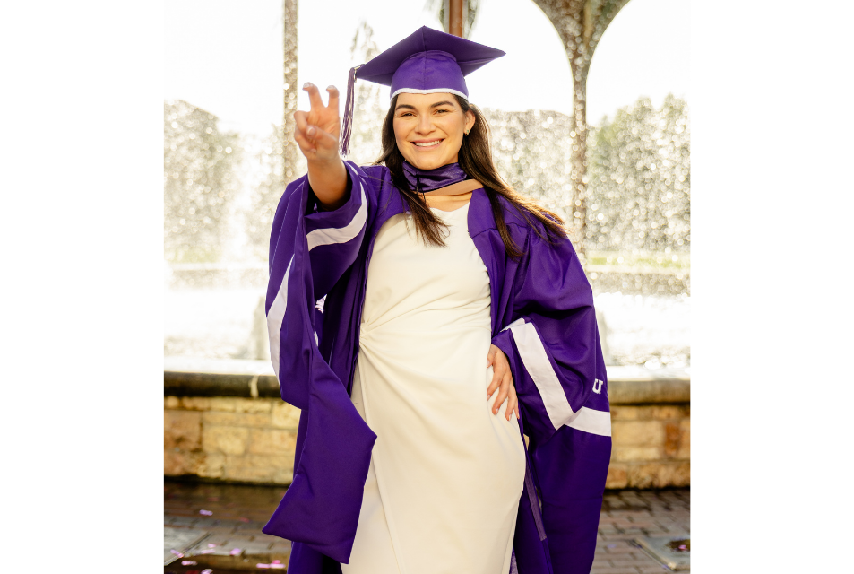 Graduate student wears her regalia and displays Horned Frog hand signal in front of Frot Fountain.